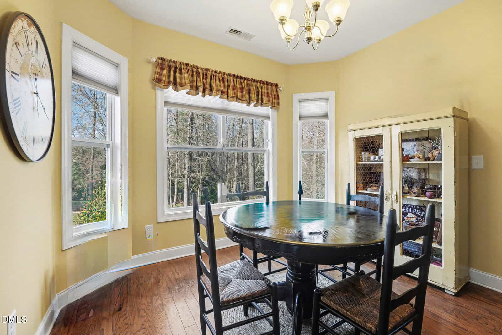 Sunny dining area with a round dark wood table, four chairs, a glass-fronted cabinet with decor, large windows with wooded view, clock on wall, hardwood floor, chandelier, and yellow walls.