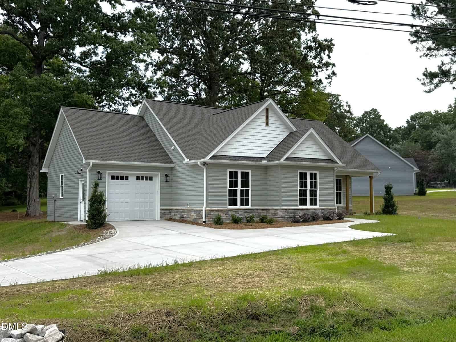 A single-story house with gray siding, white trim, and a front porch. It has a one-car garage, a concrete driveway, and a lawn with small shrubs. Trees and another house are in the background.