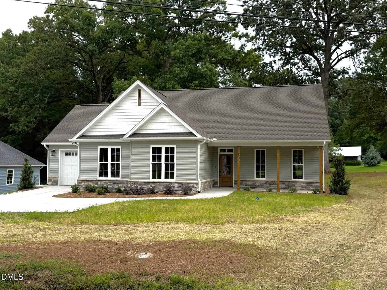 A gray single-story house with a gable roof, attached garage, front porch with wooden columns, and large windows is situated on a grassy lot with trees in the background. This charming residence is located at 306 S Elm Street in Creedmoor.