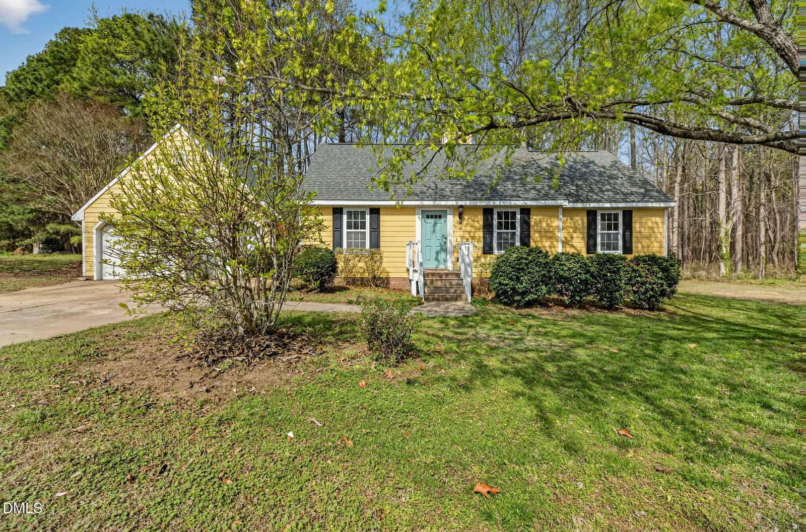 A yellow single-story house with white trim, a teal front door, and a front porch. A detached garage is on the left, and the yard has grass, shrubs, and large trees with green leaves.