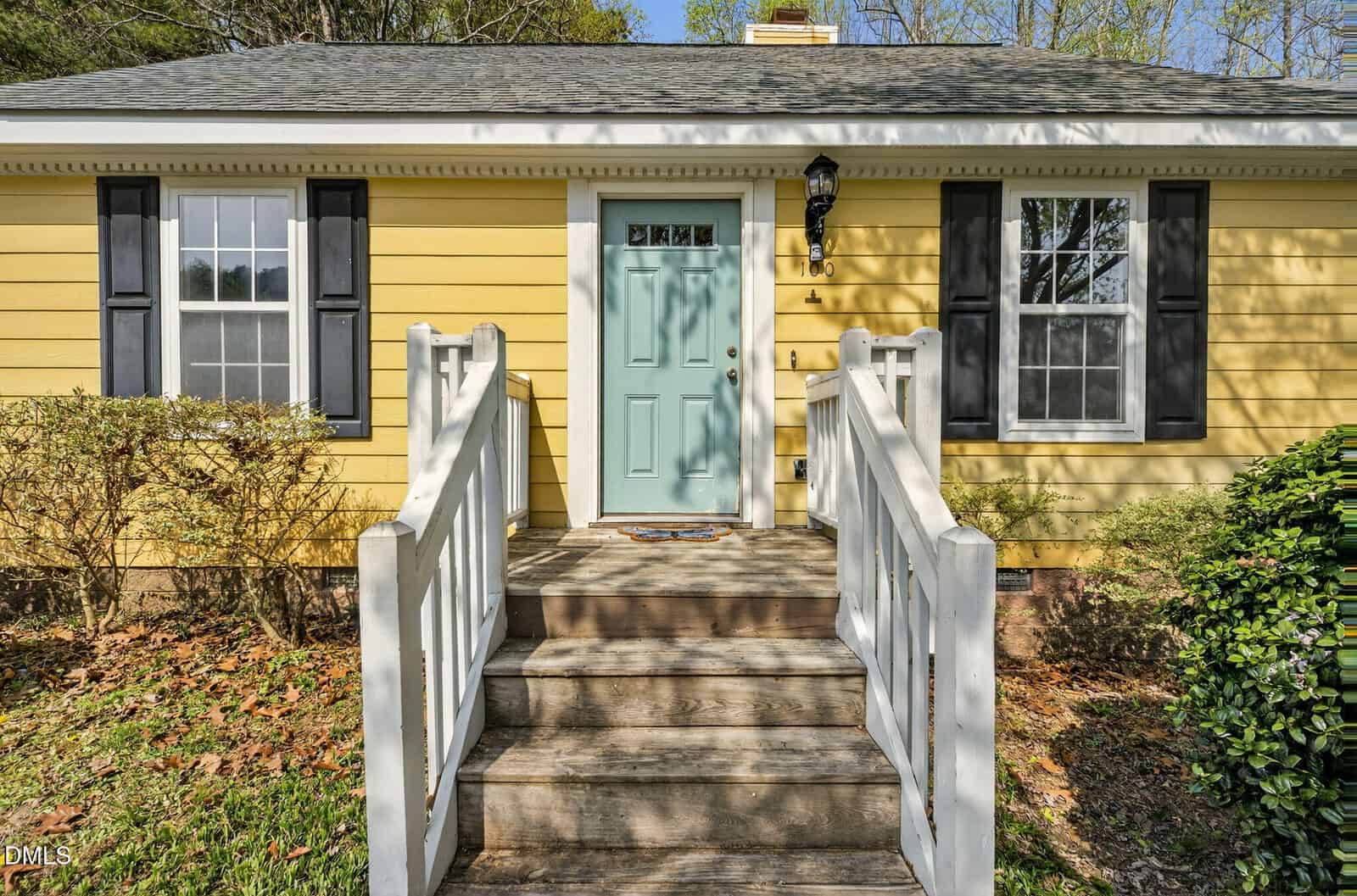 A yellow house with white trim and black shutters, featuring a green front door. A set of wooden steps with white railings leads up to the entrance, and shrubs are visible on either side of the steps.