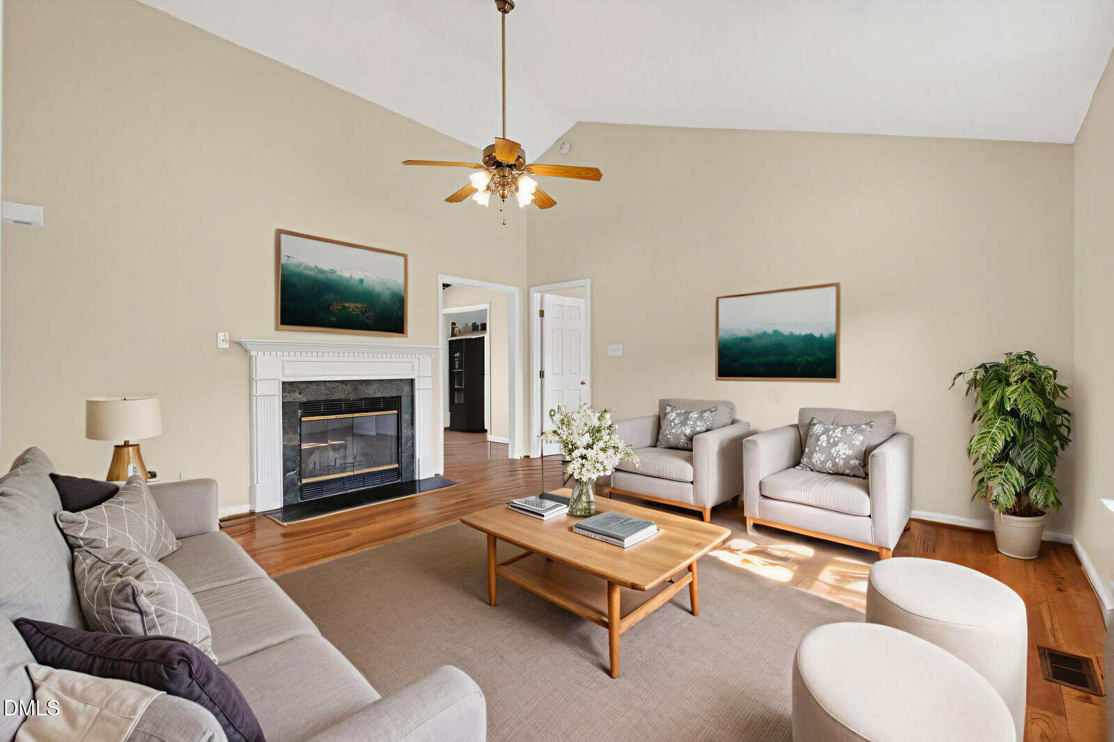 A living room with a beige wall, gray sofa, two armchairs, a fireplace with a white mantel, a wooden coffee table, a floor lamp, a potted plant, and two landscape paintings on the wall.