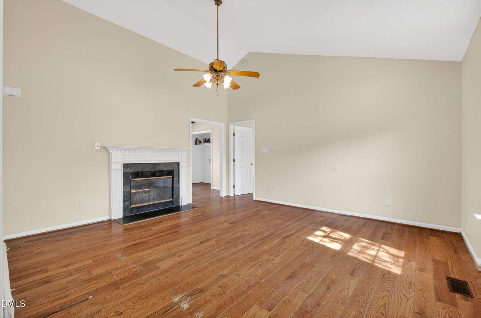 A spacious living room with hardwood floors, a white fireplace, a ceiling fan with lights, beige walls, and a high ceiling. Two doorways lead to other rooms, and natural light enters through a window.