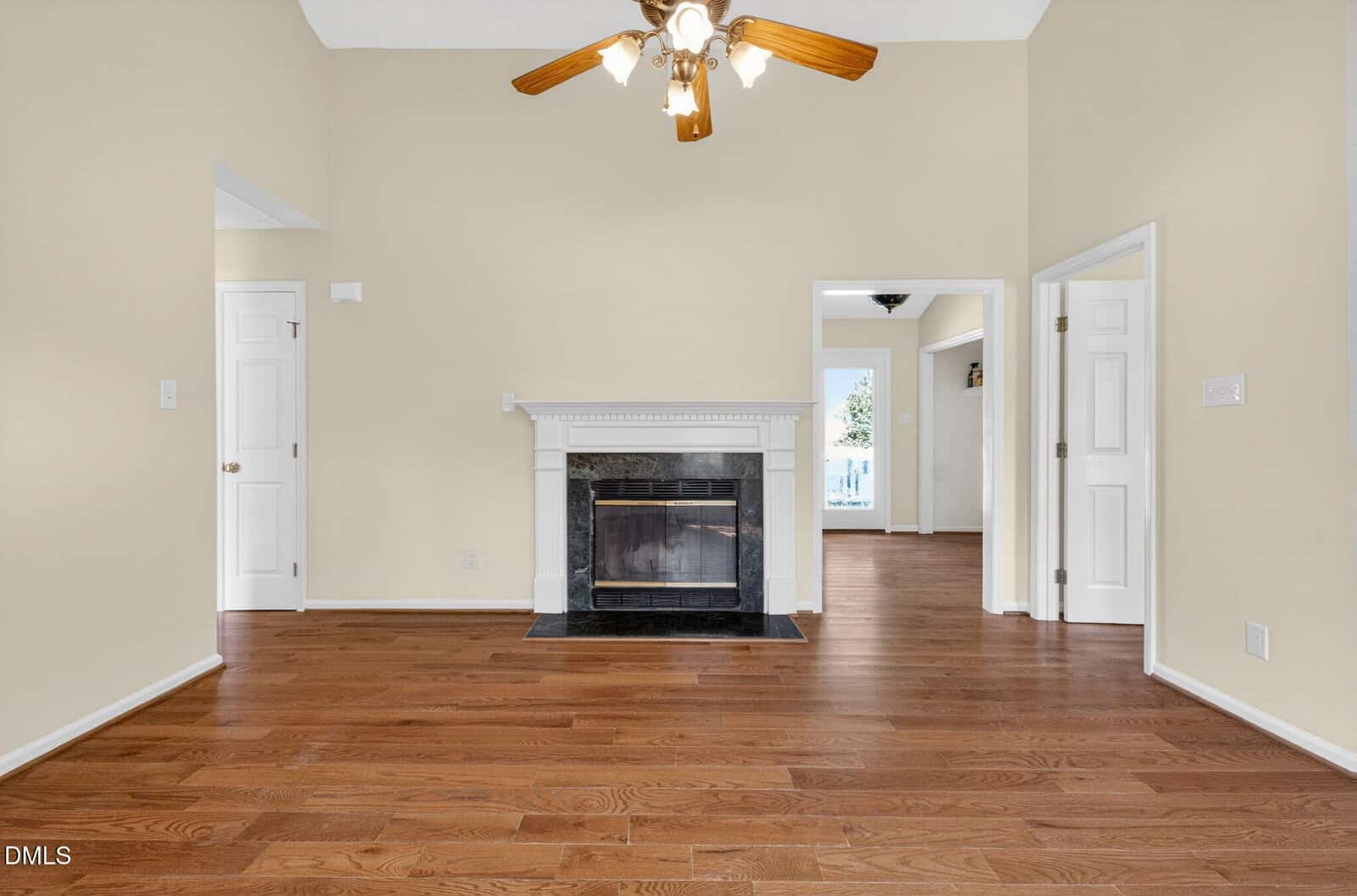 Living room with wood floors, beige walls, a white fireplace, and a ceiling fan with five wooden blades. Three doors lead to other rooms, and natural light comes from a window in the adjacent room.