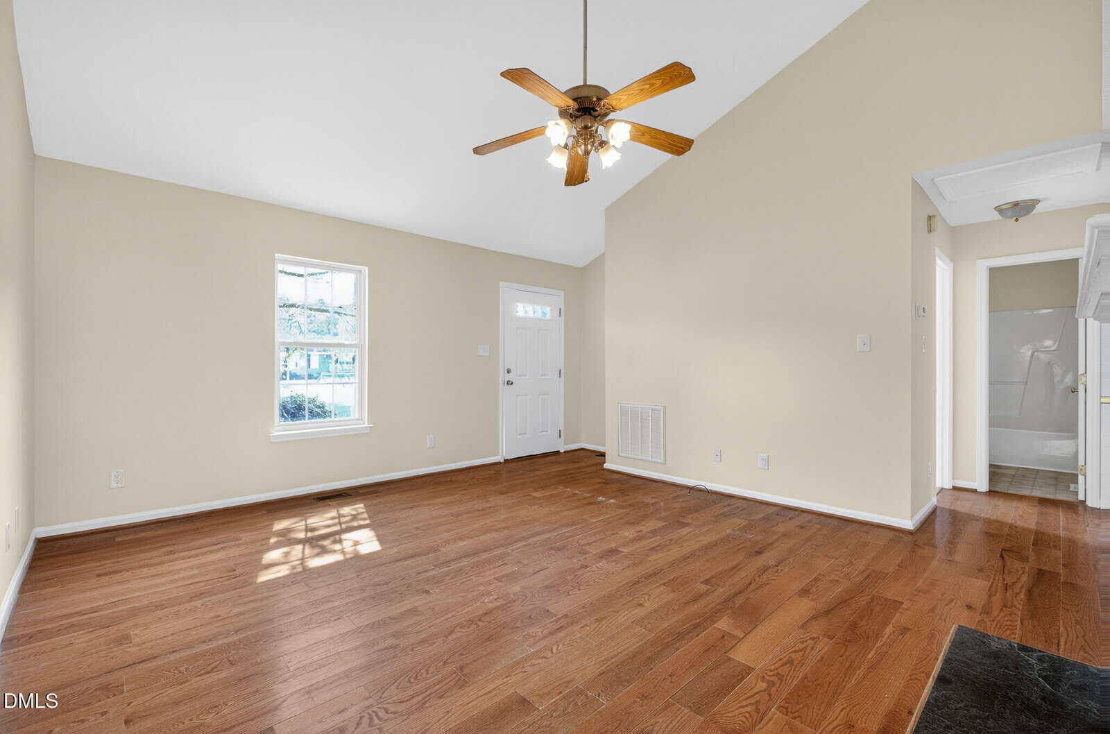 A bright, empty living room with hardwood floors, beige walls, a ceiling fan with lights, two windows, and a white front door. A hallway leads to another area of the house.