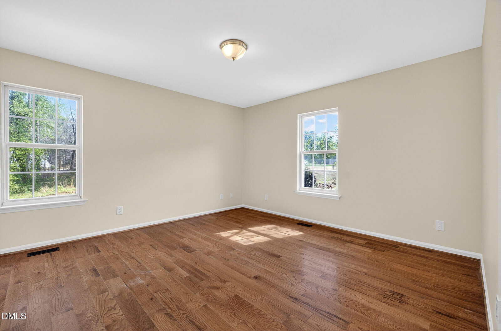 Empty room with beige walls, two large windows letting in natural light, a ceiling light fixture, and polished wooden floors. Greenery and blue sky are visible through the windows.