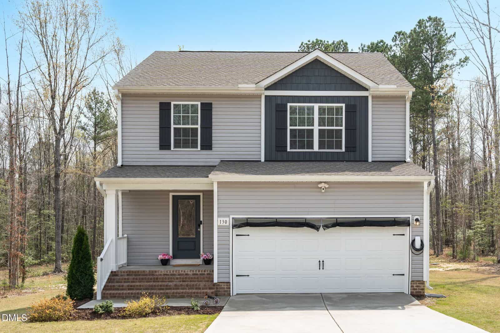 A two-story suburban house with gray siding, black shutters, and a front porch. The house has a double garage and is surrounded by a lawn with some small shrubs, set against a backdrop of trees.