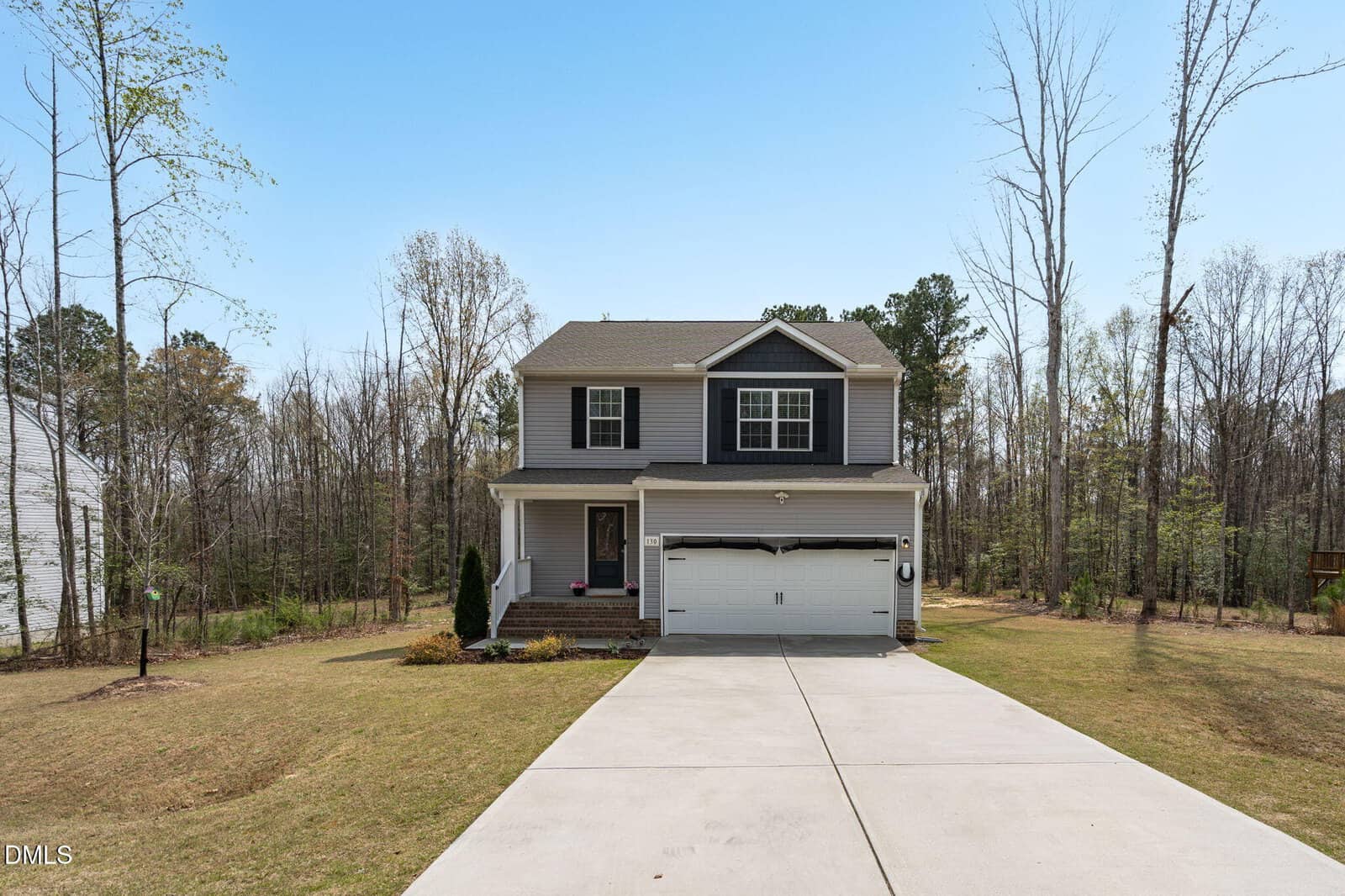 A two-story house with gray siding, black shutters, and a white double garage sits at the end of a concrete driveway. The lawn is bordered by tall trees and the sky is clear.