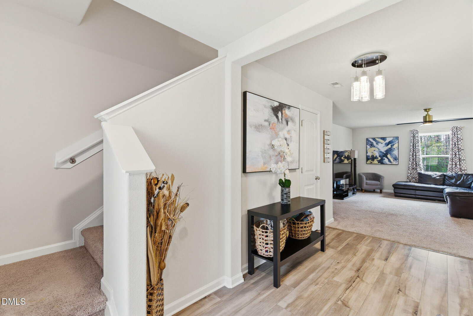 Modern home entryway with wood flooring, a black console table holding a vase of flowers and baskets, stairs to the left, and a bright living room with sofas and large windows in the background.