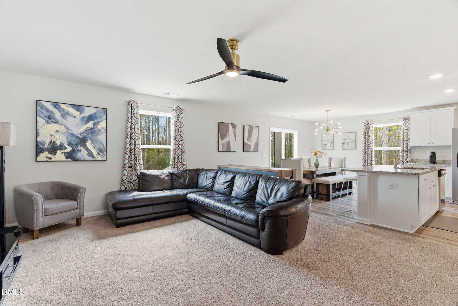 A living room with a black leather sectional sofa, gray armchair, abstract wall art, carpeted floor, and large windows with patterned curtains. The space opens to a dining area and kitchen with white cabinetry.
