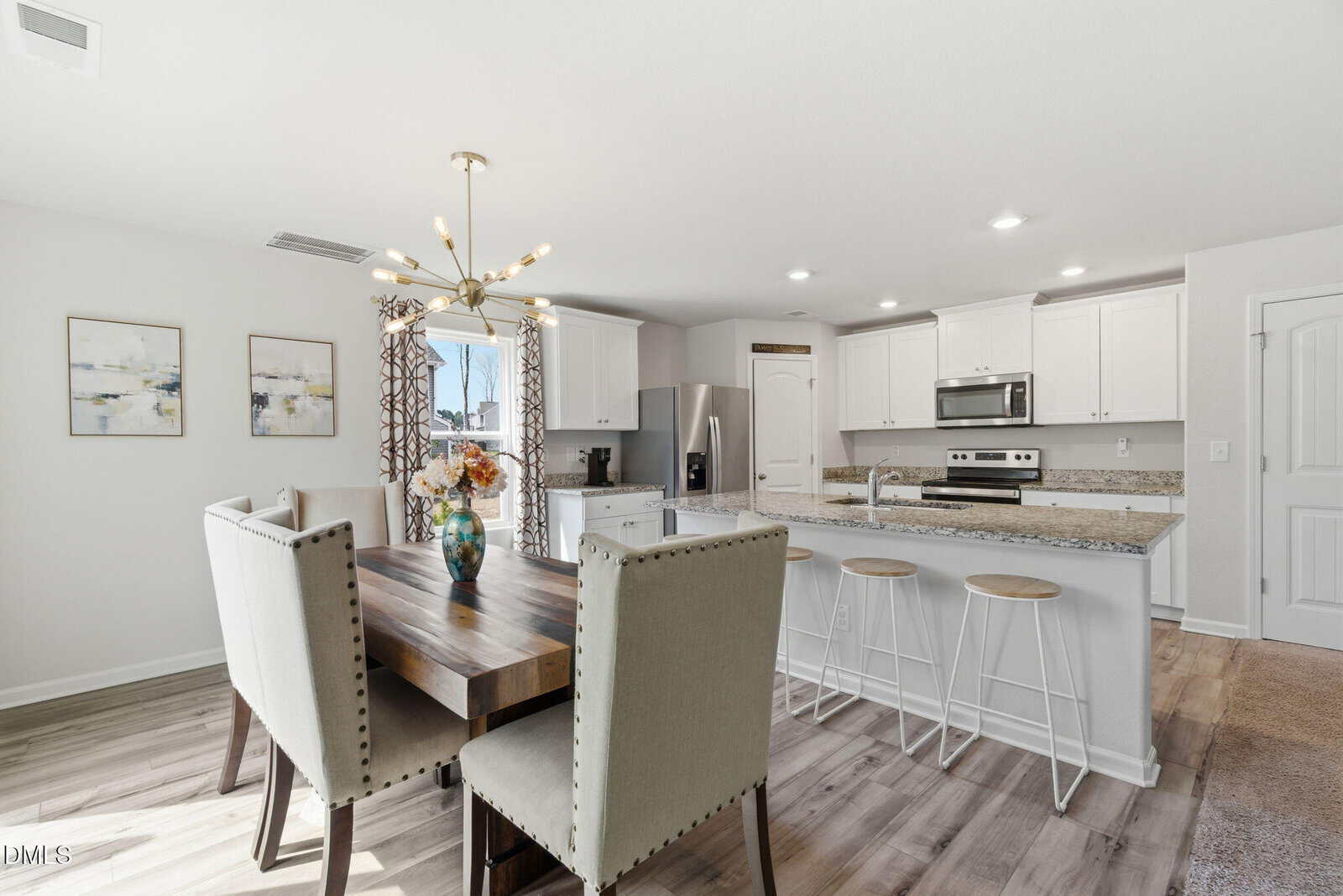 Bright kitchen and dining area with a wooden table, upholstered chairs, granite countertops, white cabinets, and barstools. Sunlight streams through a glass door with curtains; modern chandelier and wall art are visible.