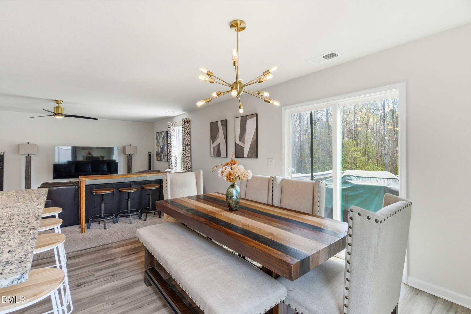 A modern dining area with a striped wood table, upholstered chairs, and a bench is shown next to large windows overlooking a wooded area. A gold chandelier hangs above, and a kitchen island and living room are visible in the background.