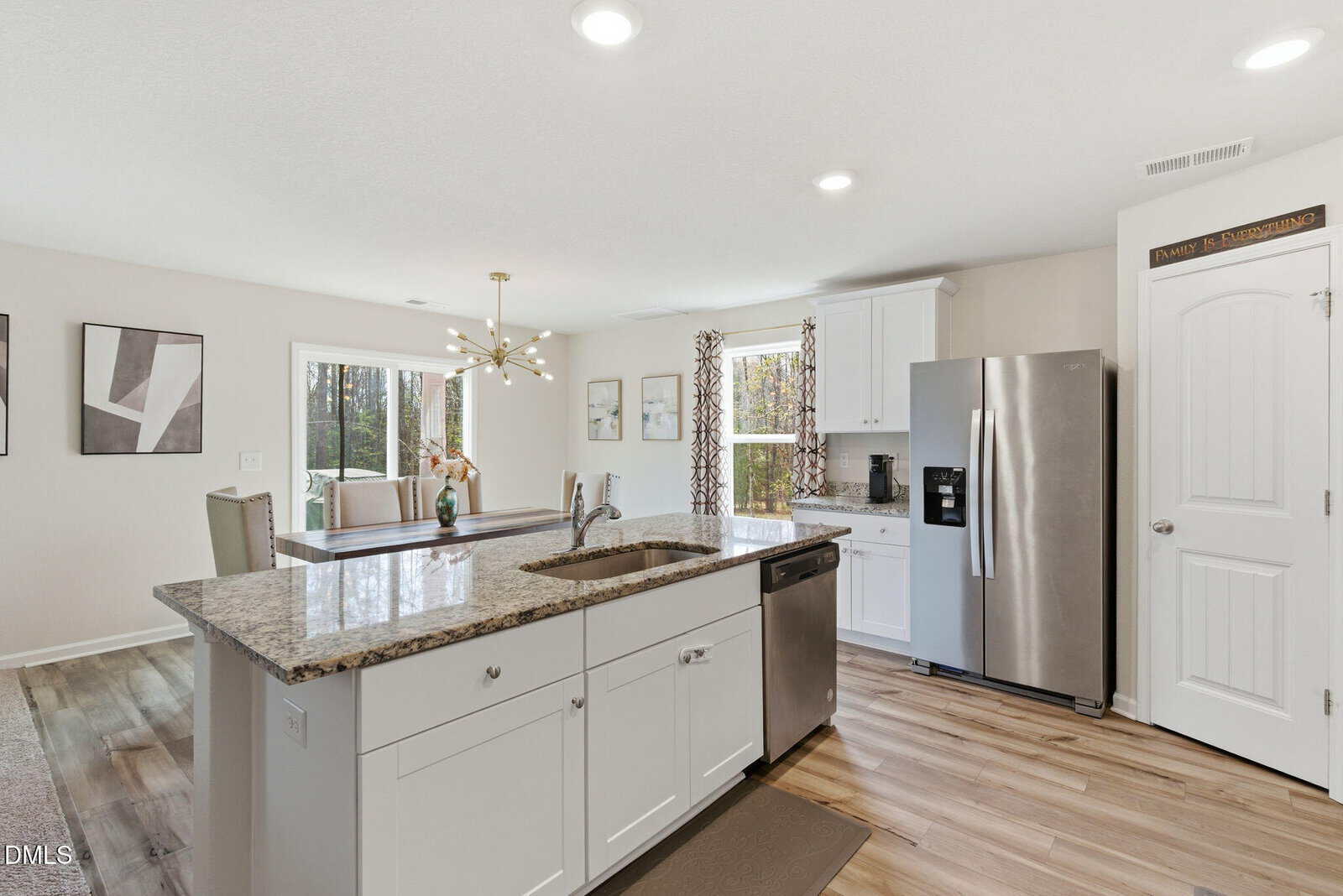 Modern kitchen with white cabinets, stainless steel appliances, granite island with sink, light wood flooring, and a view into a dining area with windows and wall art. Bright, natural light fills the space.