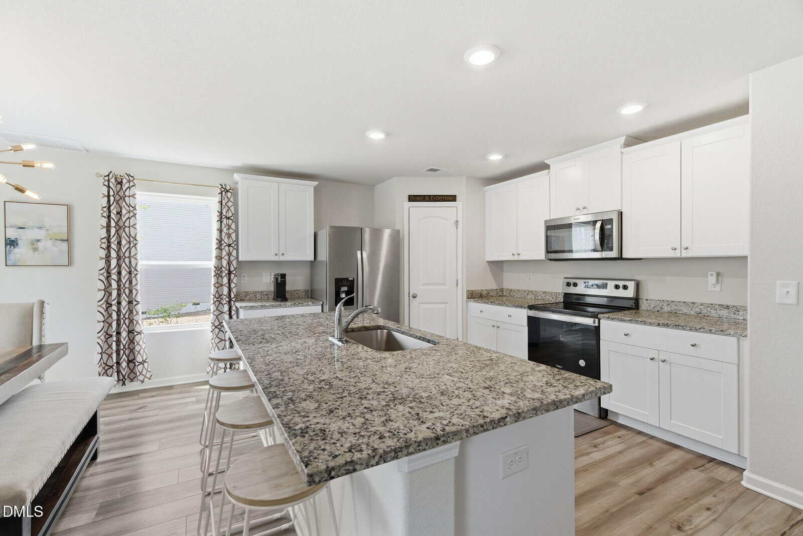 Modern kitchen with white cabinets, stainless steel appliances, granite countertops, a center island with a sink and barstools, wood-style flooring, and a window with patterned curtains letting in natural light.