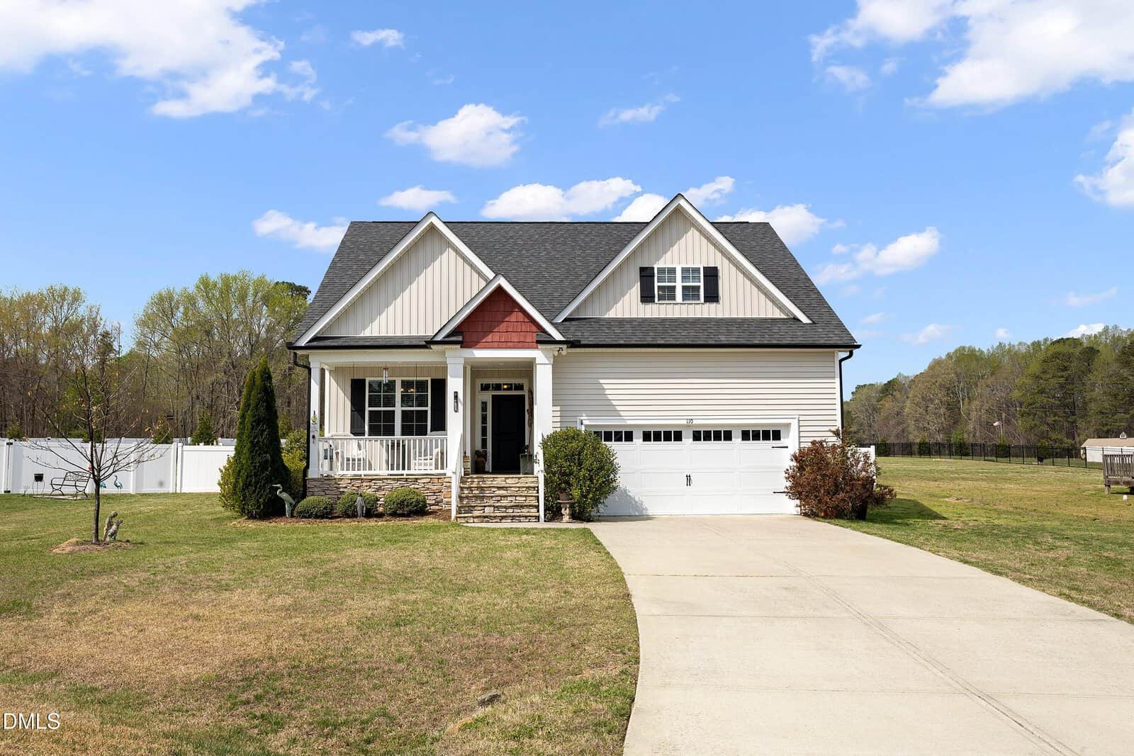 A single-family house with white siding, dark roof shingles, a front porch, attached garage, and a concrete driveway. The house is surrounded by grass and trees under a blue sky with scattered clouds.