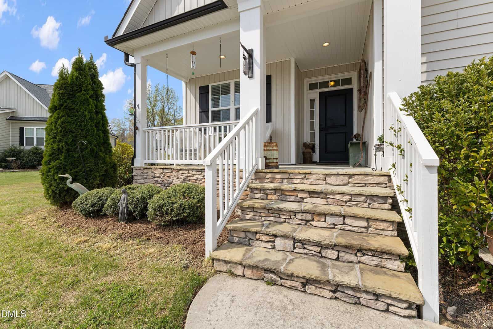 A stone staircase with white railings leads up to a covered porch with columns. The house has light siding, a black front door, and landscaping with bushes and a small lawn. The sky is blue with scattered clouds.