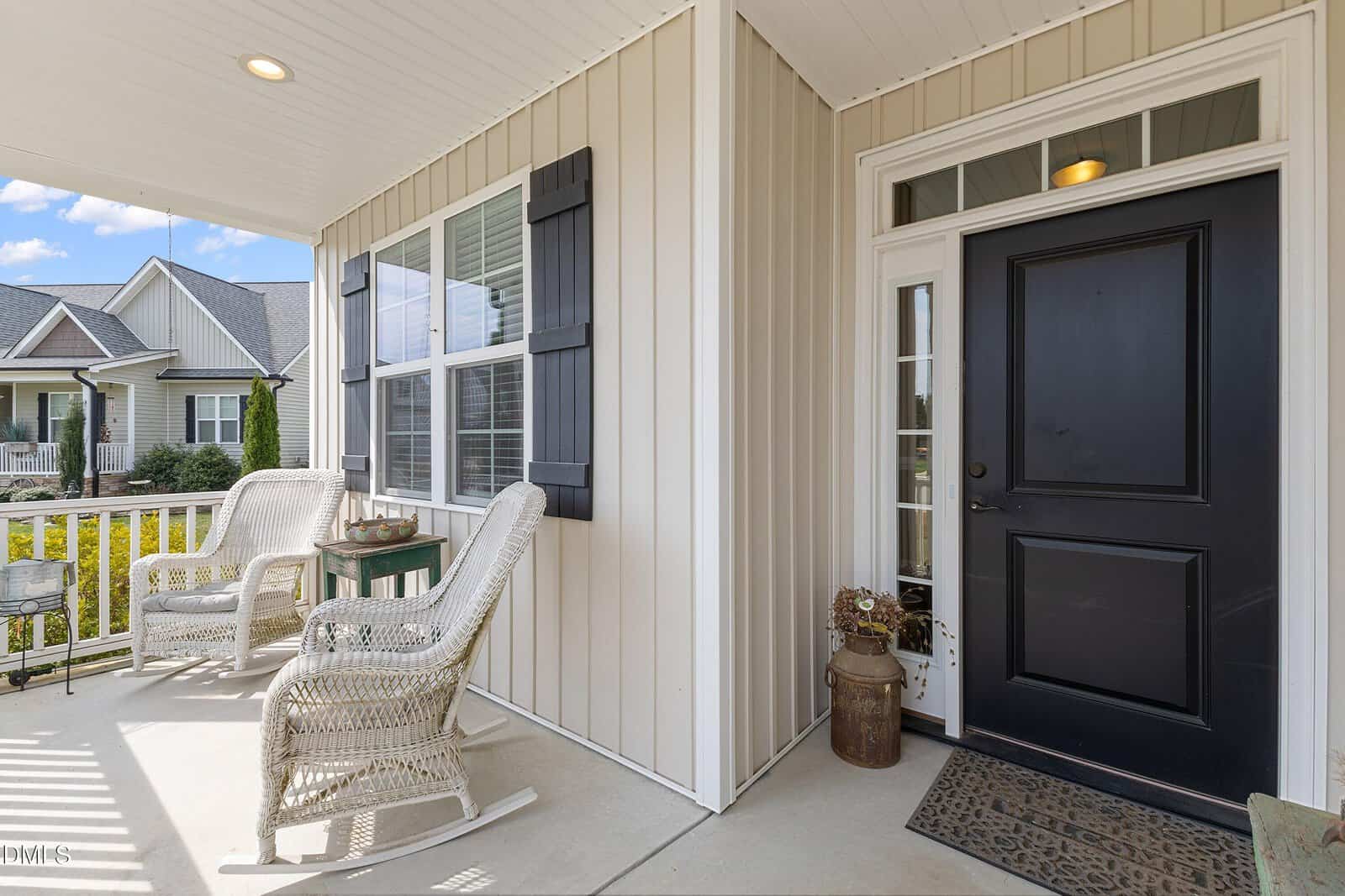 Covered front porch with two white wicker chairs, a small side table, light-colored siding, black shutters, and a black front door with sidelights. A milk can and doormat are positioned near the door.