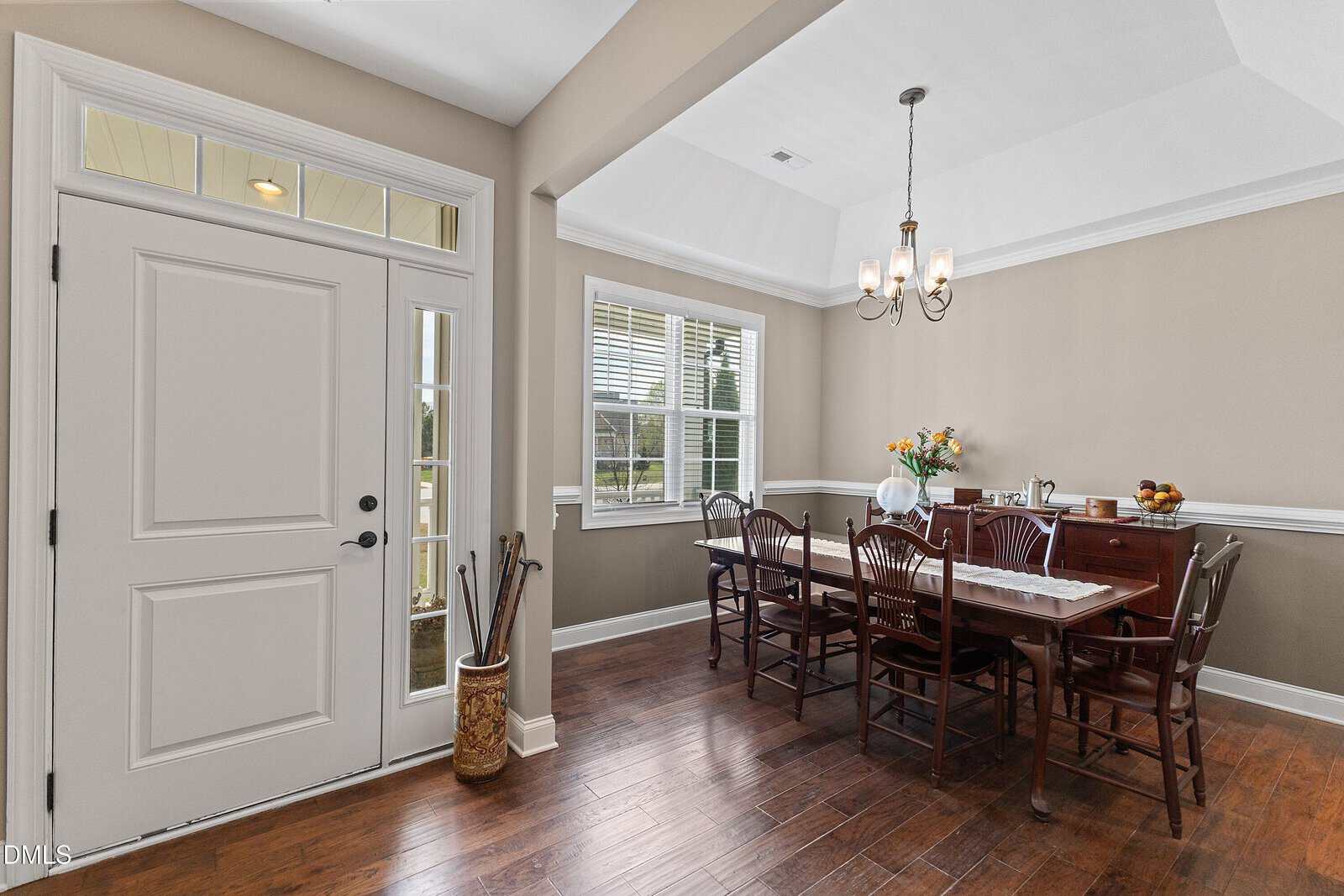 A dining room with a rectangular wooden table and six chairs, a chandelier overhead, and a window letting in natural light. A sideboard with flowers and decor sits against the wall. The entry door is visible on the left.