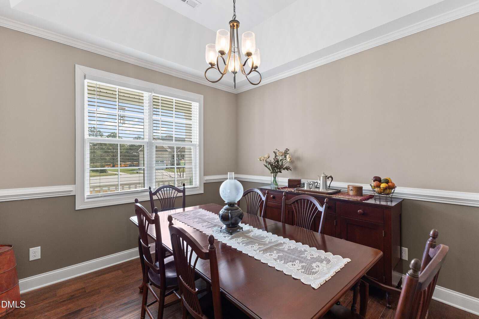A dining room with a wooden table and six chairs, a lace table runner, a blue glass lamp, a sideboard with decor items, beige walls, white trim, and sunlight coming through a window.