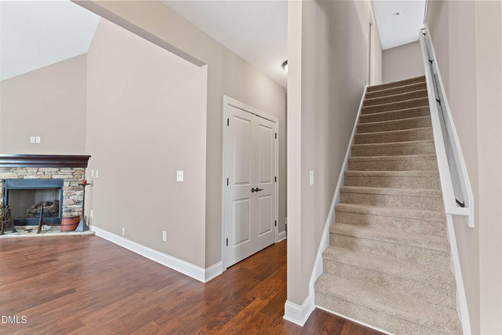 Carpeted stairs with white railing lead up next to a hallway with double doors. To the left is a stone fireplace and hardwood floor in a room with beige walls and white trim.