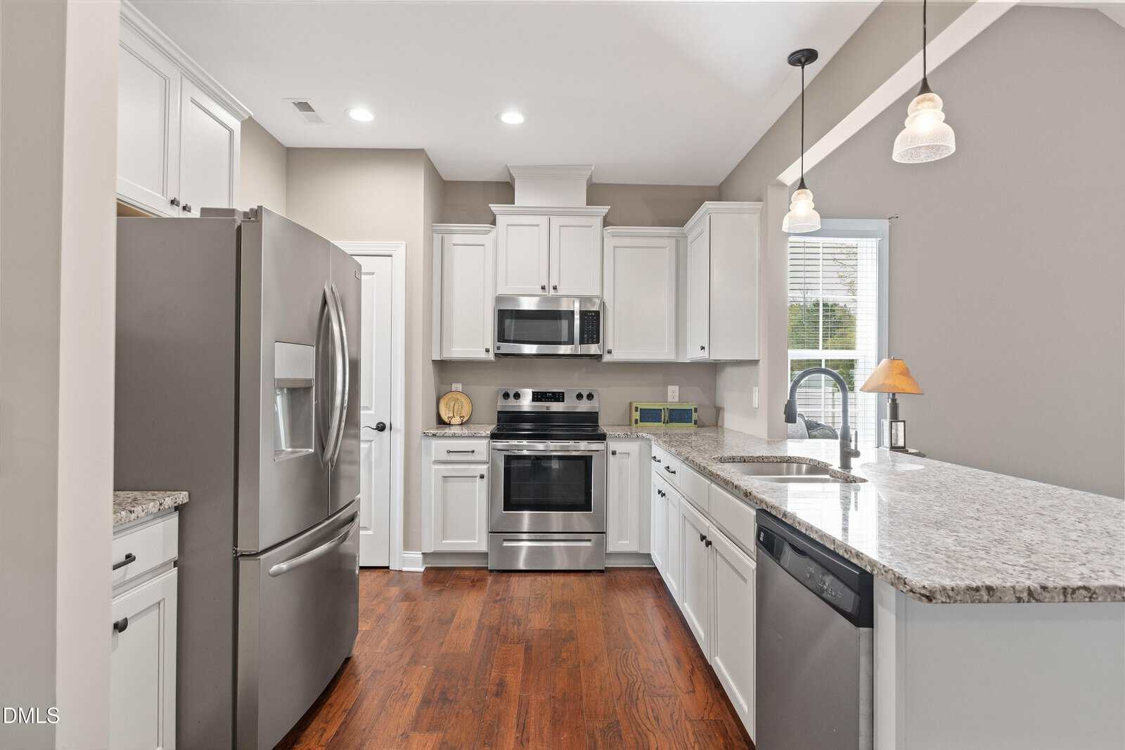 Modern kitchen with stainless steel appliances, white cabinets, granite countertops, a large refrigerator, microwave, stove, dishwasher, and pendant lights above the sink. Hardwood floor and a window add warmth and light to the room.