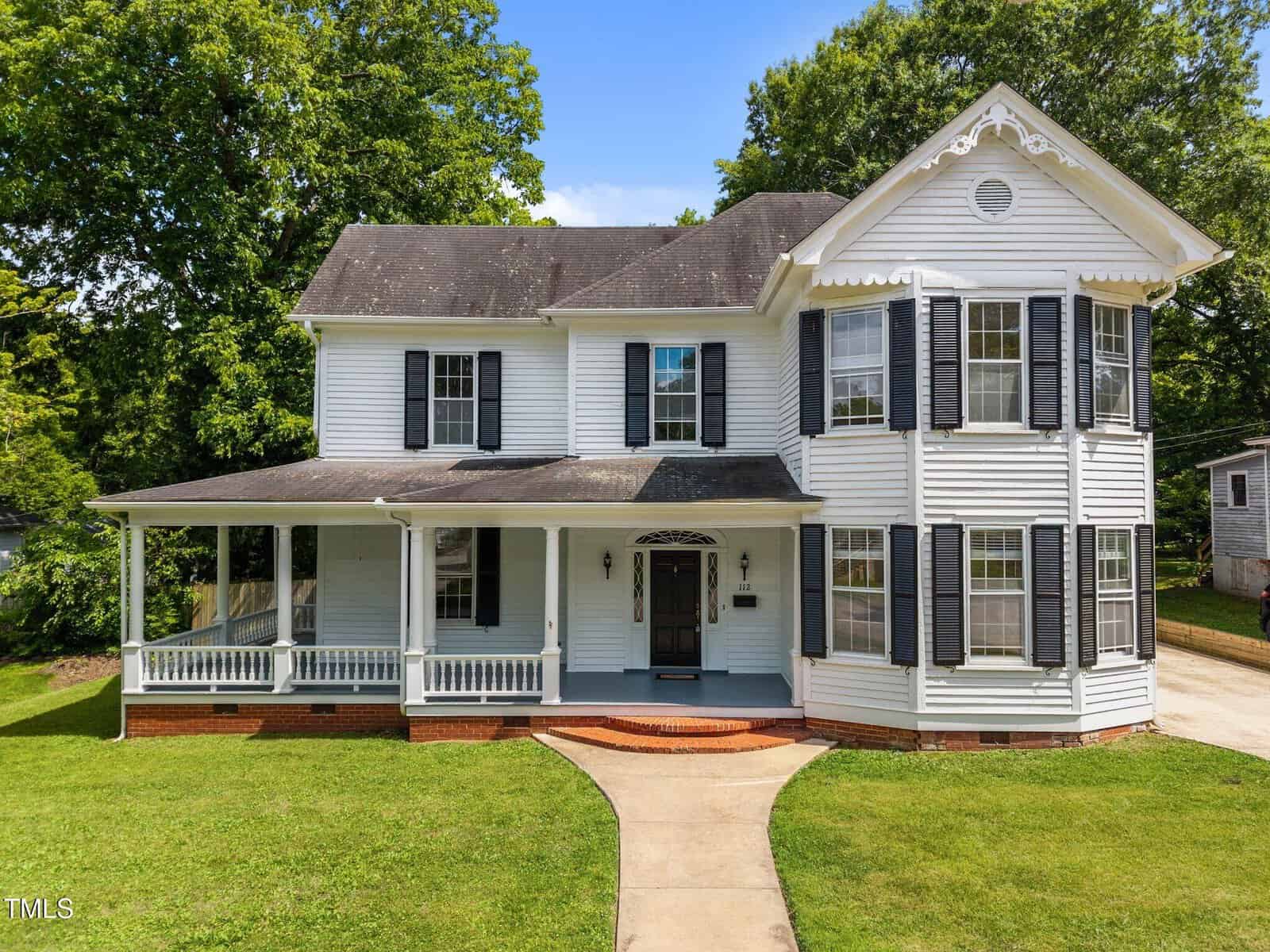 Two-story white house with black shutters and a covered front porch. The house has a prominent bay window section on the right, a well-kept lawn, and a curved brick walkway leading to the front entrance.