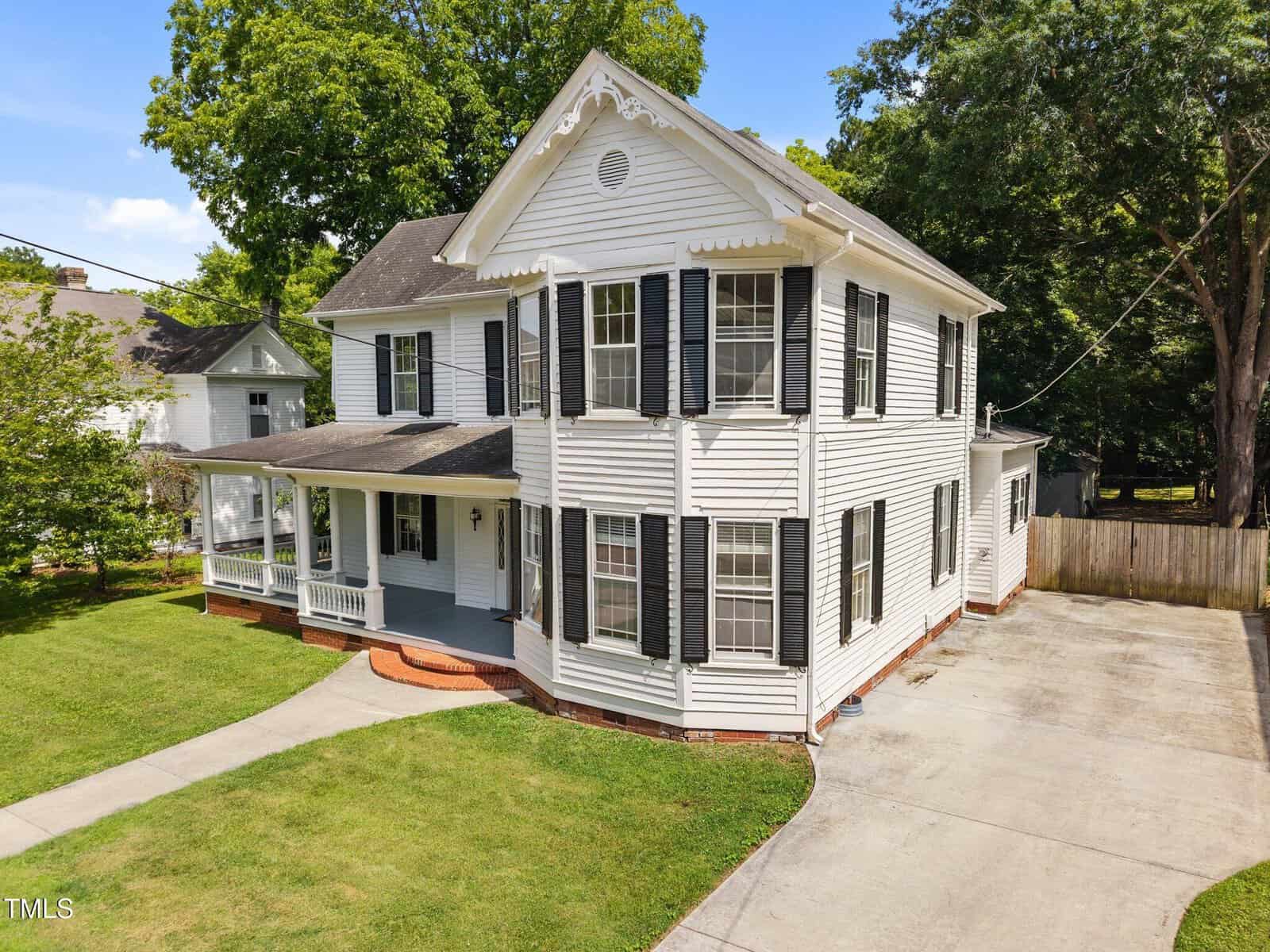 A two-story white house with black shutters, a covered front porch, and a bay window sits on a well-maintained lawn with a curved driveway and a fenced backyard. Trees are visible in the background.