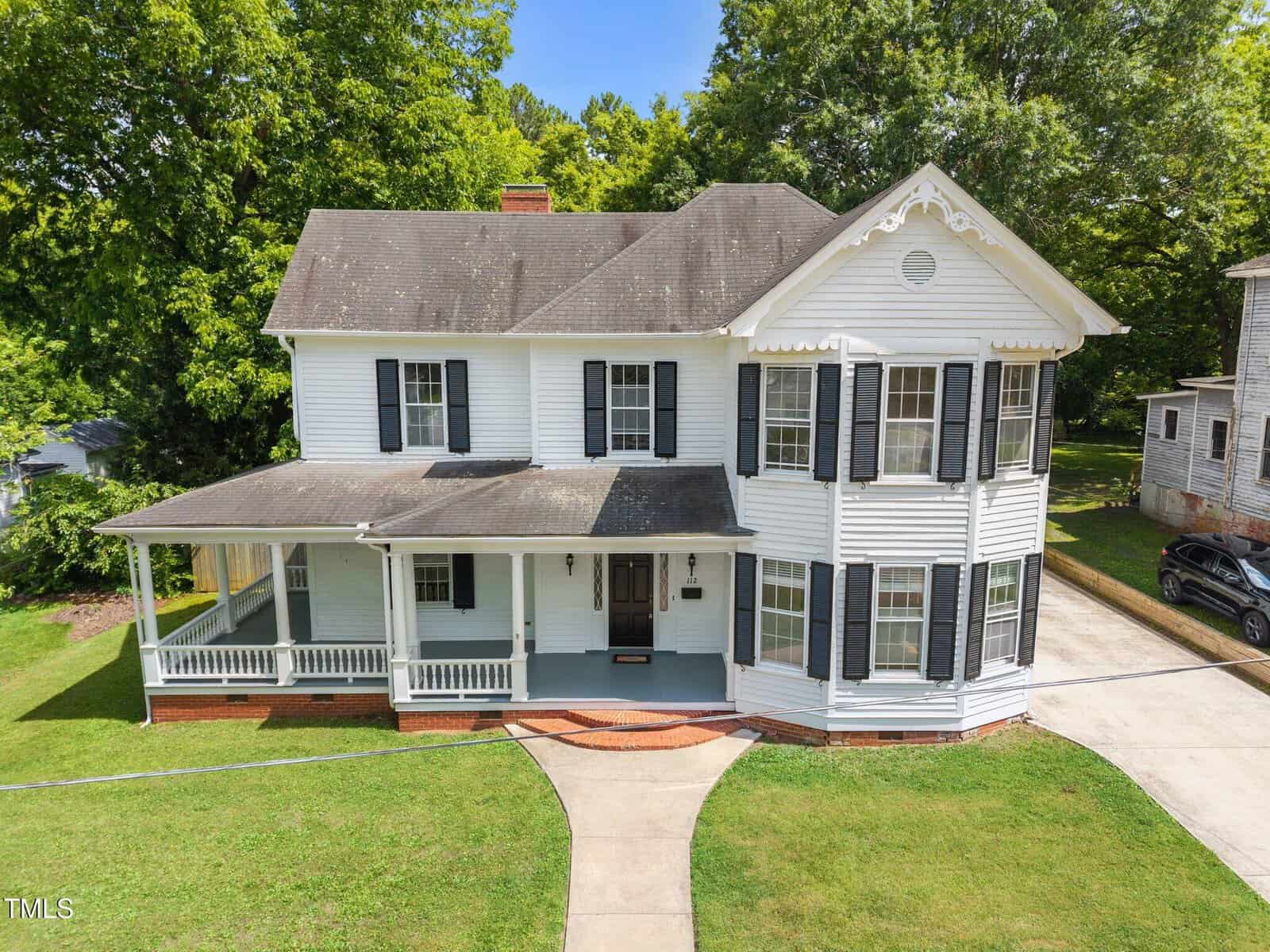 A two-story white house with black shutters and a covered front porch. The house has a gabled roof, decorative trim, and sits on a well-kept lawn with a curved walkway leading to the front door.