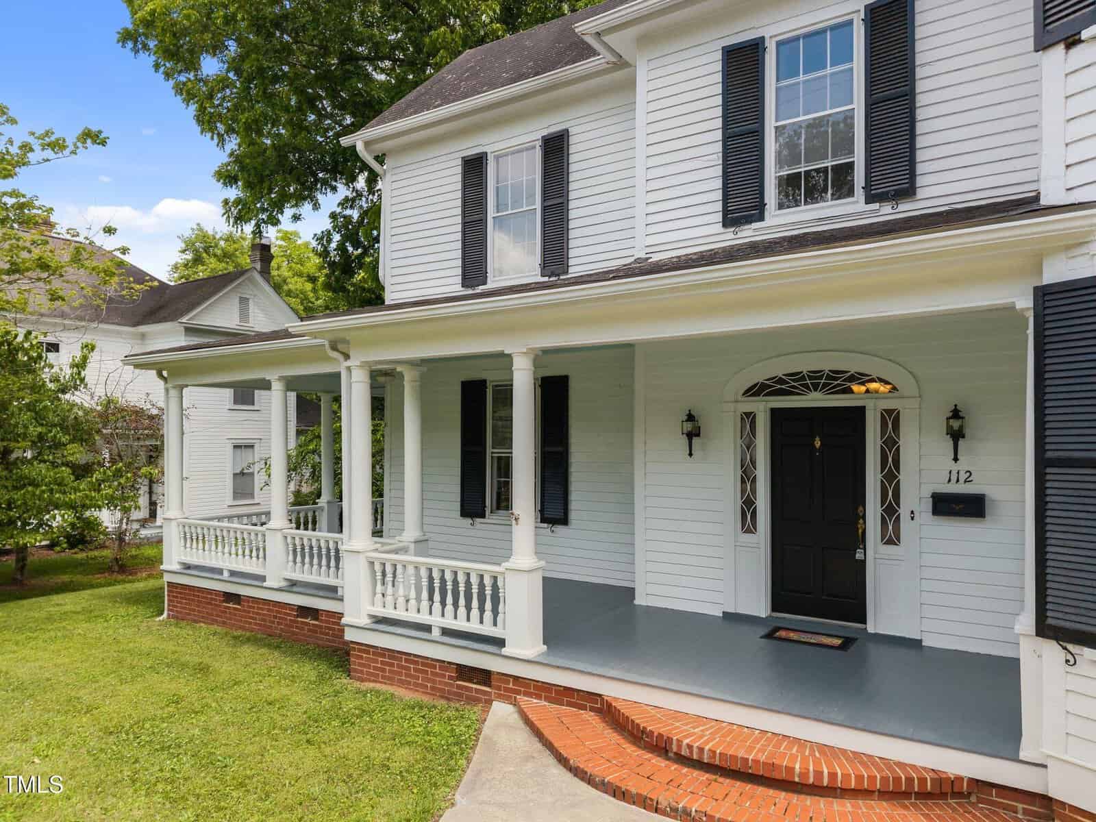 A two-story white house with black shutters, a dark front door, and a wide covered porch with white railings and columns. The porch is accessed by brick steps and the yard has green grass and trees.