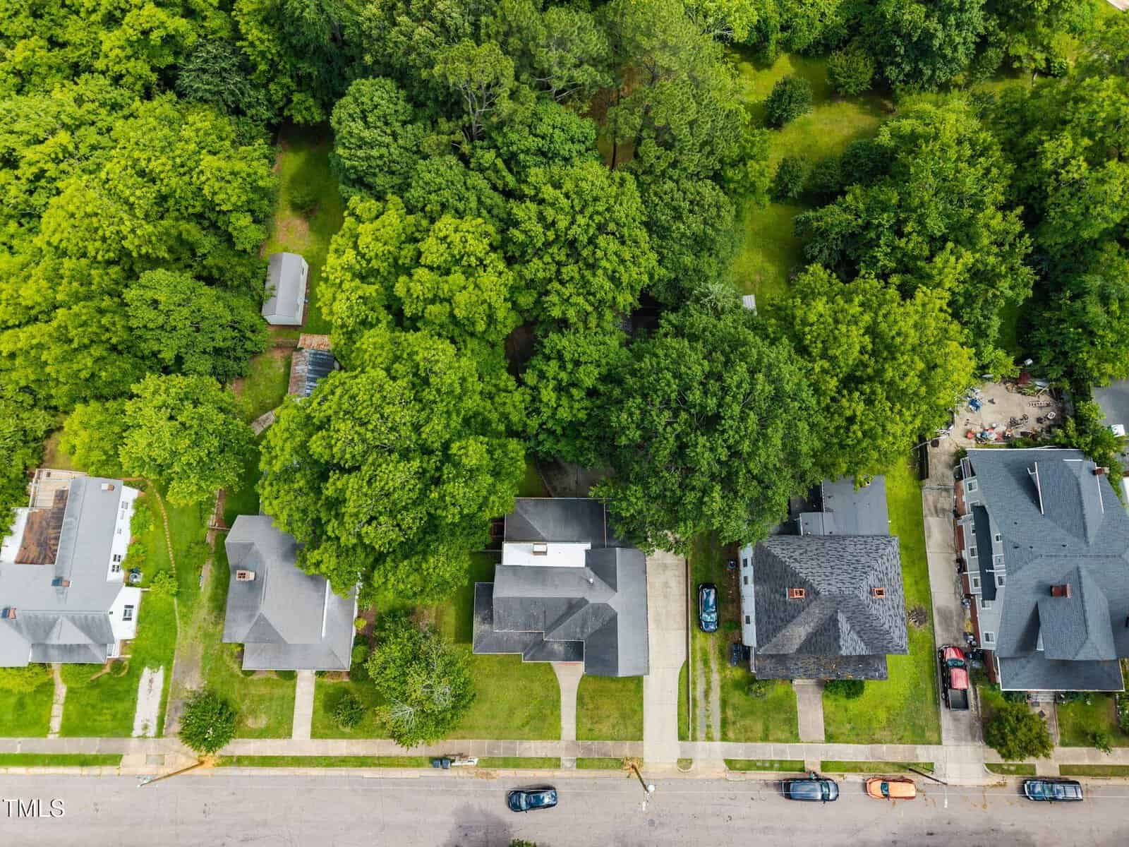 Aerial view of a suburban neighborhood with houses, driveways, parked cars, lawns, and abundant green trees filling backyards and surrounding the area.