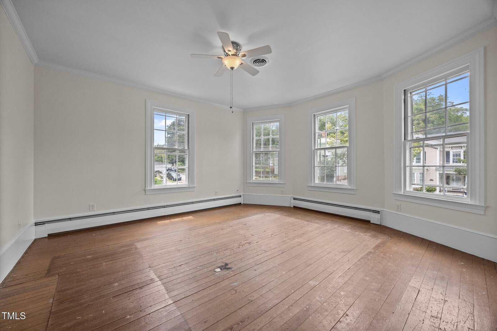 Bright, empty room with four large windows, a ceiling fan, and light beige walls. The hardwood floor appears worn, and there is baseboard heating along the walls. Trees and houses are visible through the windows.