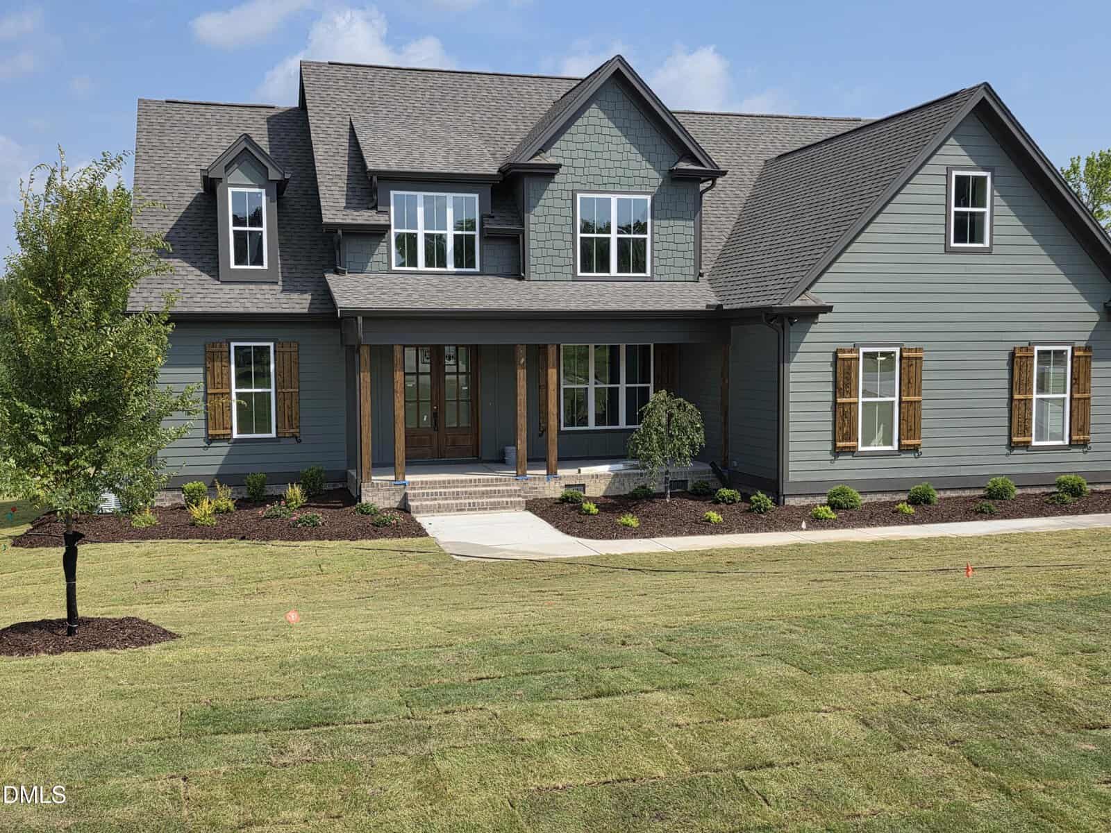 A charming two-story suburban house at 115 Water Willow Lane in Zebulon, featuring gray-green siding, wooden shutters, a covered front porch, and a newly landscaped lawn with fresh sod and small shrubs.