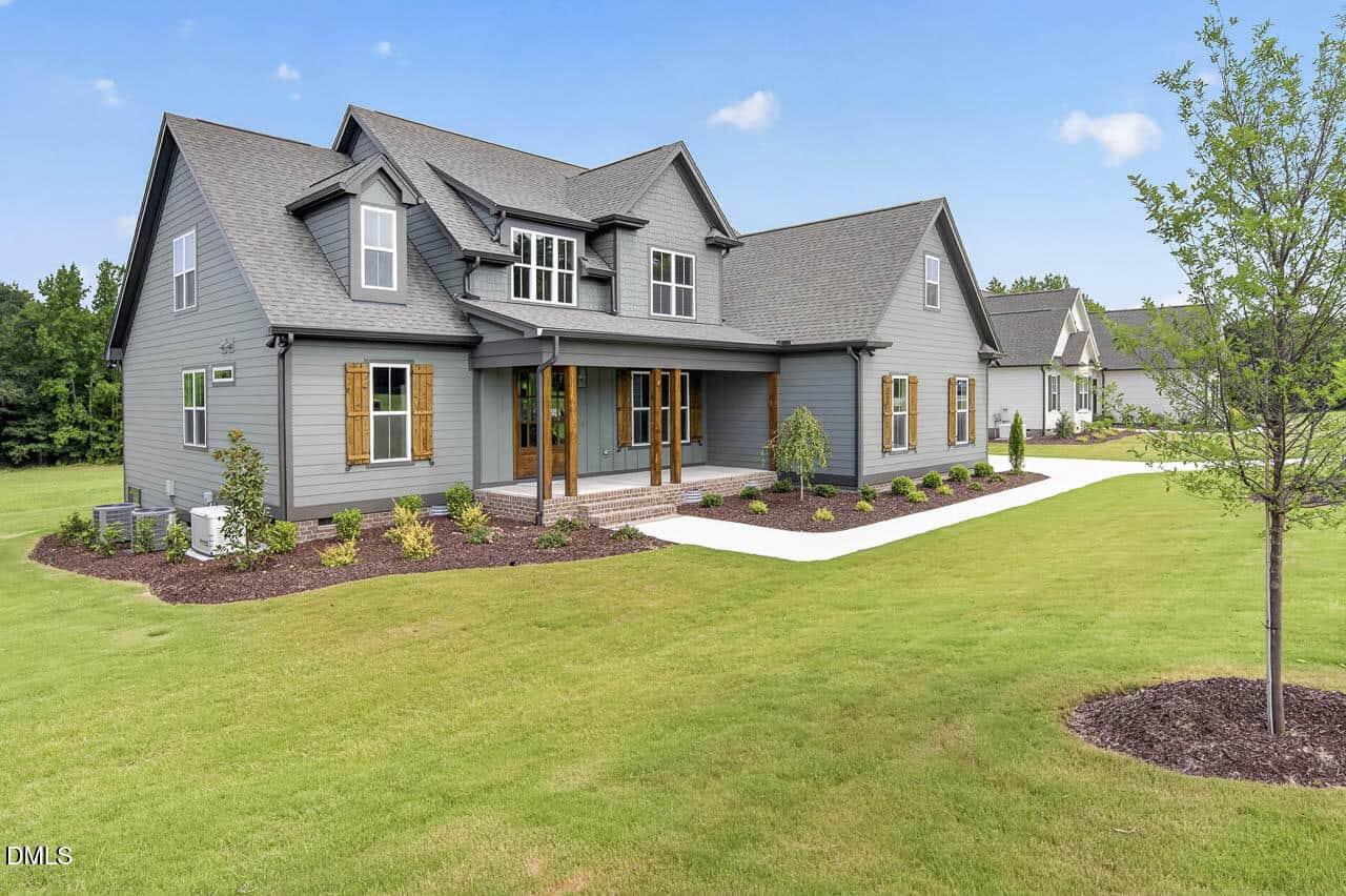 Gray two-story house with brown shutters, large front porch, manicured lawn, landscaped garden beds, and a tree in the foreground under a clear blue sky.