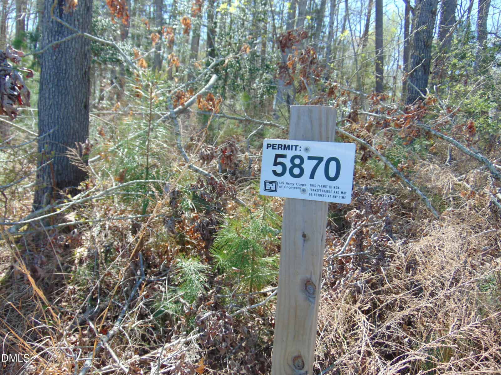 A wooden post with a sign reading "PERMIT: 5870" stands in front of dense, leaf-covered underbrush and trees in a wooded area on a sunny day.