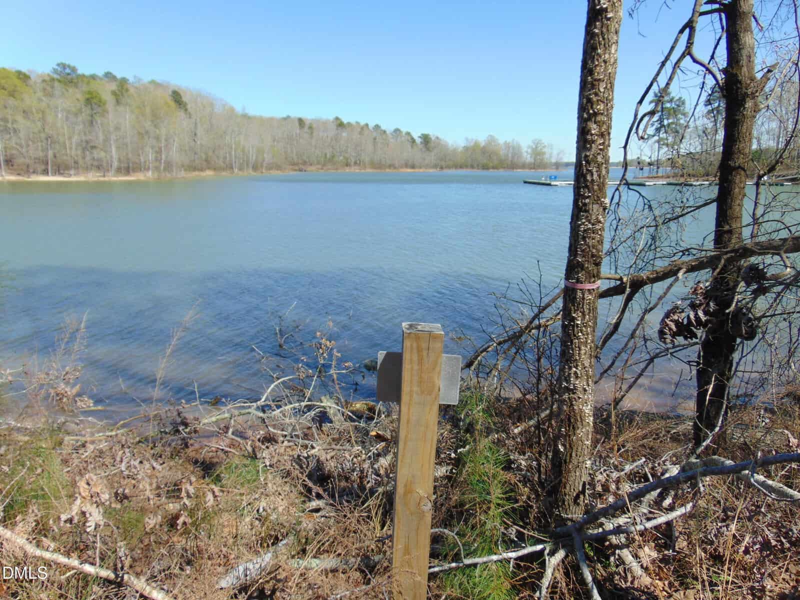 A wooden post stands near the edge of a calm lake with clear blue water, surrounded by leafless trees and a forested shoreline under a clear sky. A small boat is visible in the distance on the water.