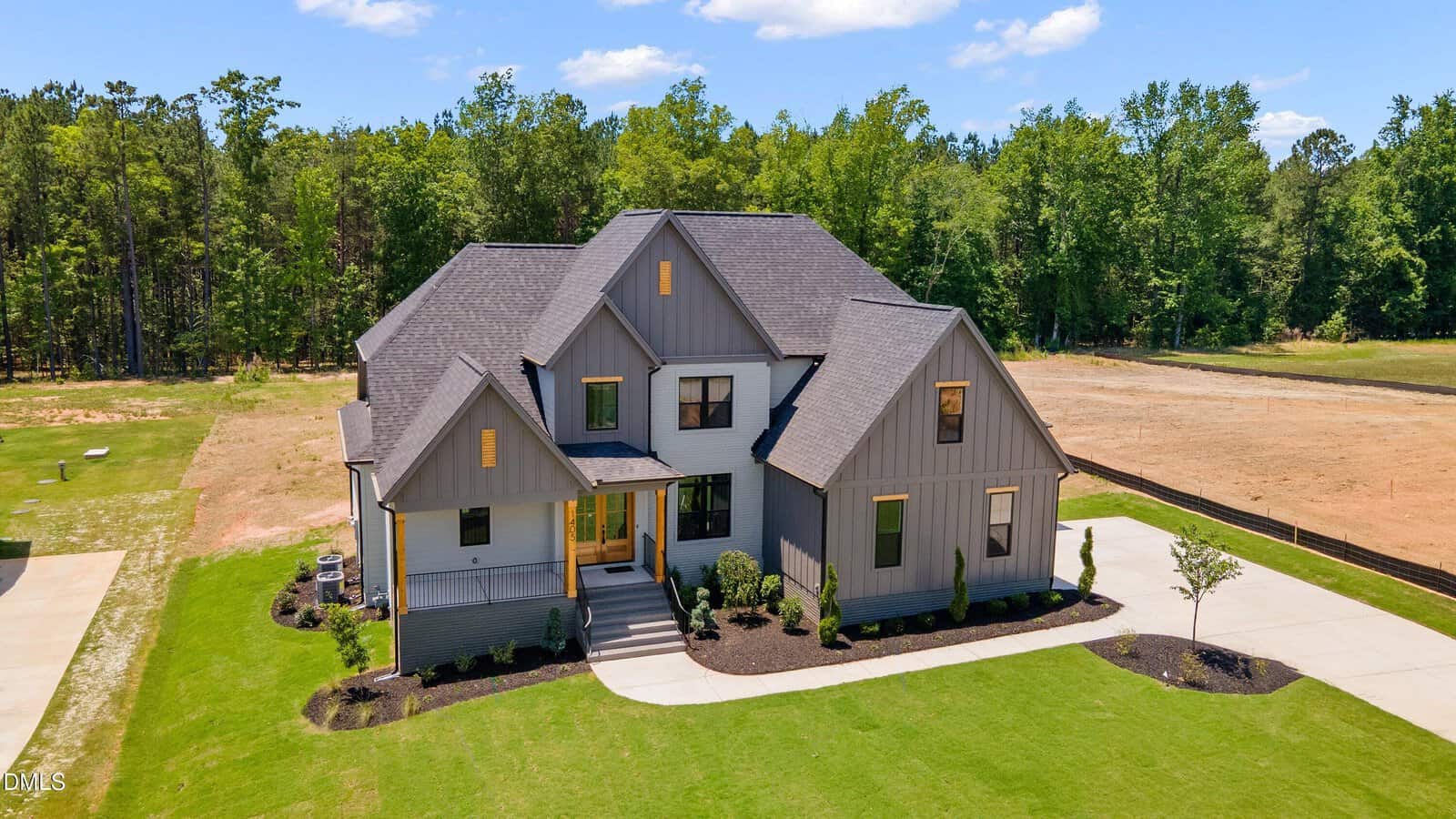 A large modern farmhouse with gray siding, multiple gables, and a well-maintained lawn sits beside a wooded area under a blue sky at 1405 Cottondale Lane in Wake Forest. The driveway curves to the front entryway.