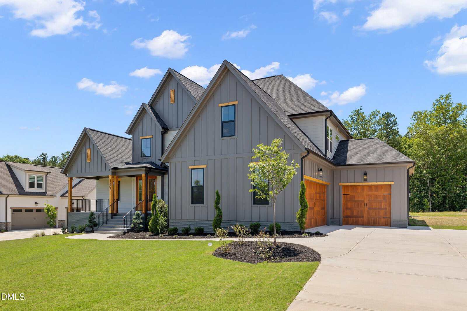 Modern two-story gray house with wooden accents, large windows, and a front porch. Neatly landscaped yard with green grass, a small tree, and a wide driveway leading to a wooden garage door. Blue sky with scattered clouds.