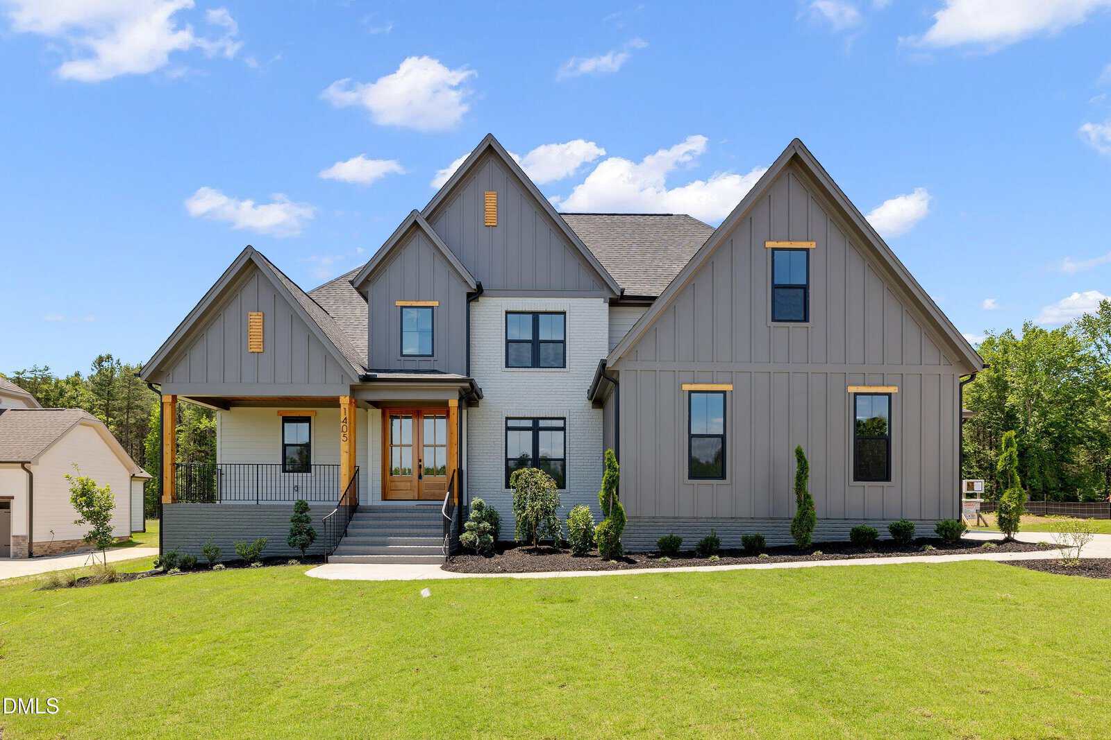 A modern two-story house with gray siding, white brick accents, and a covered front porch. The house features multiple peaked roofs, large windows, and is surrounded by a well-manicured lawn and small shrubs.