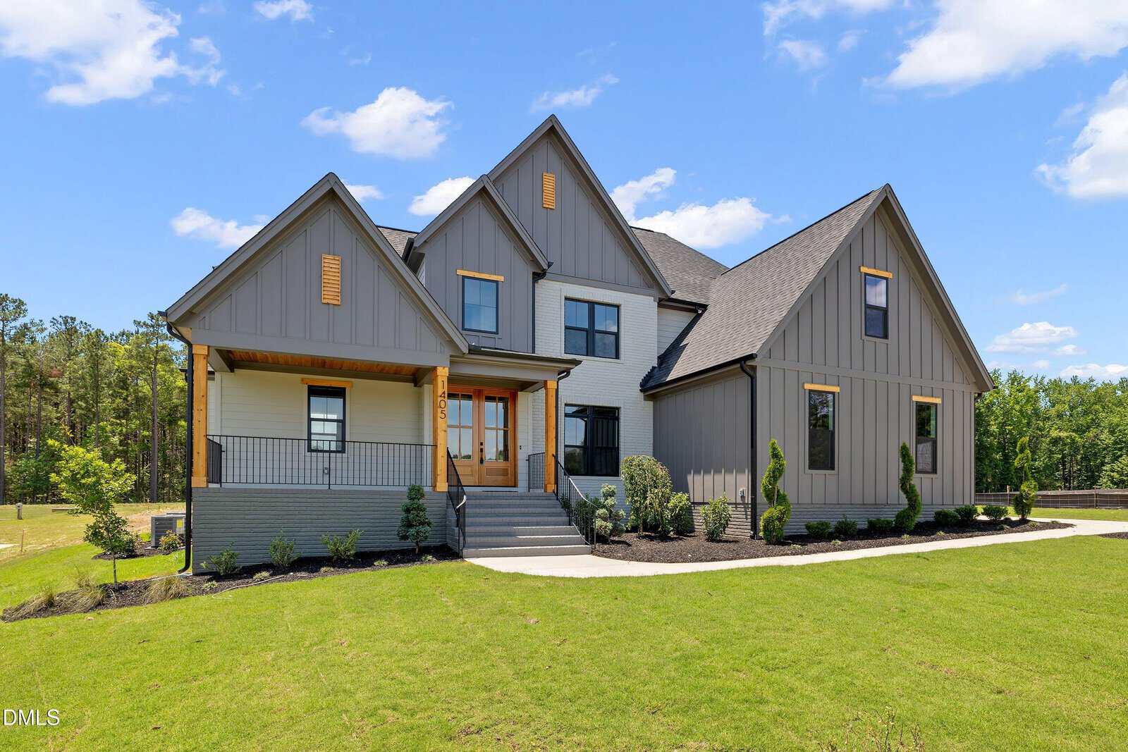 A modern two-story house with gray siding, steep gable roofs, wooden accents, a front porch with steps, landscaped yard, and a clear blue sky in the background.