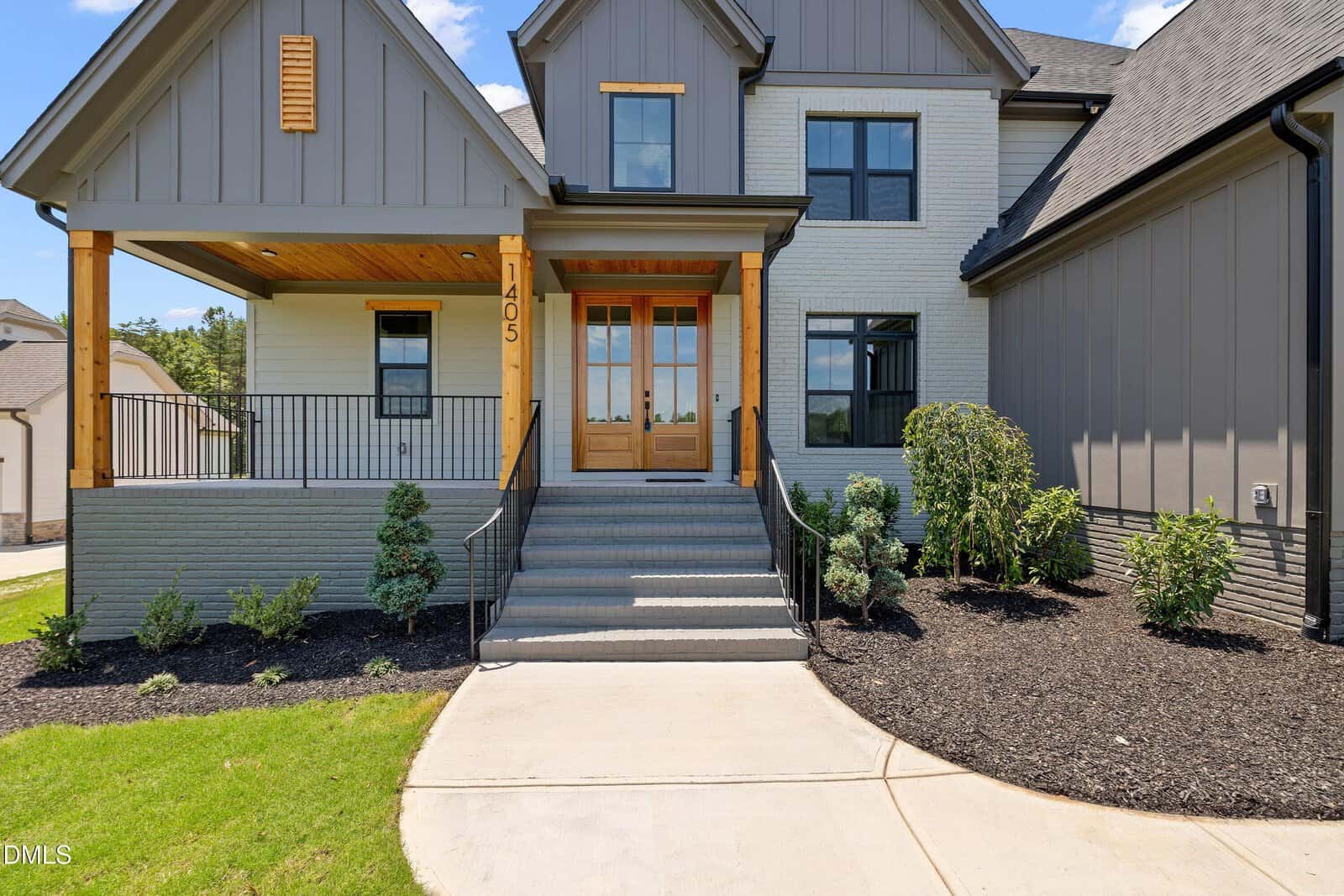 Modern two-story house with gray siding, brick accents, wooden porch posts, double front doors, large windows, landscaped yard, and a curved concrete pathway leading to the entrance.