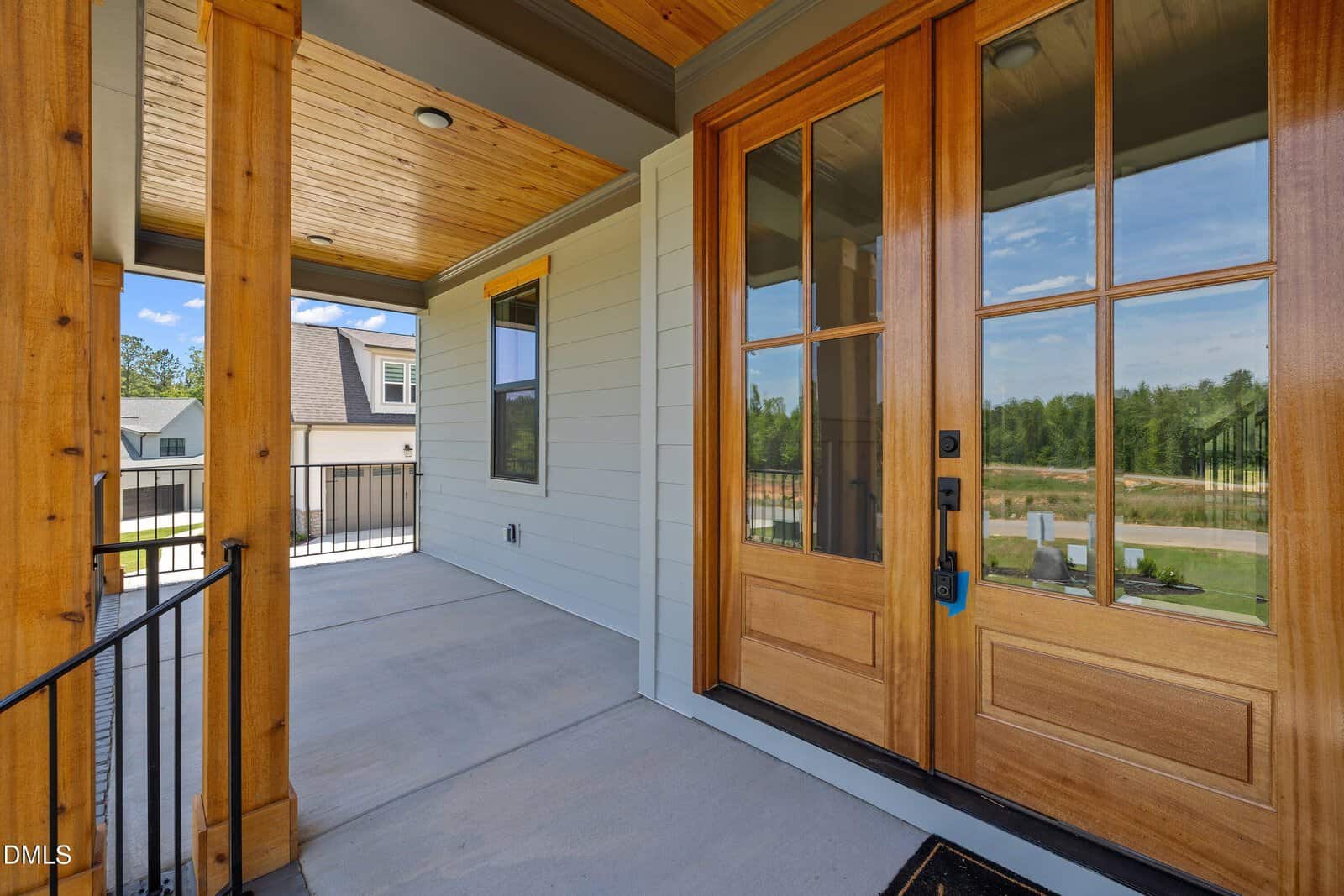 A wooden front door with glass panels opens onto a covered porch with a wooden ceiling, overlooking a suburban neighborhood and green lawns.