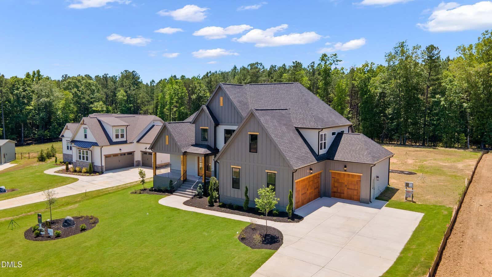 Modern two-story house with gray siding, large windows, and double wooden garage doors, set on a spacious lawn with landscaped gardens and surrounded by trees in a suburban neighborhood under a blue sky.