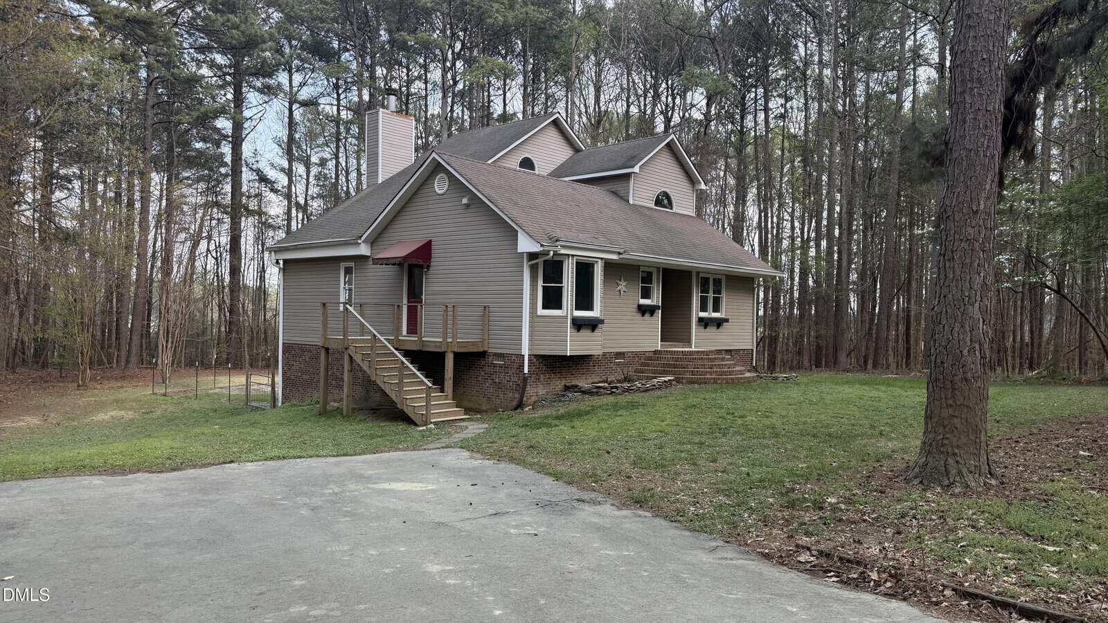 A beige, two-story house with a gabled roof, front porch with red awning, brick foundation, and elevated wooden steps, situated next to a wooded area and a paved driveway.