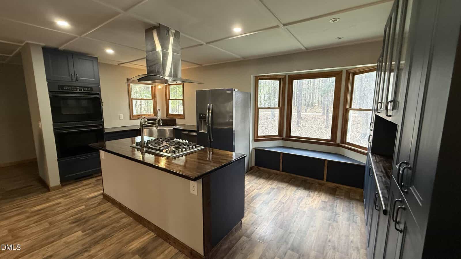 Modern kitchen with dark cabinets, stainless steel appliances, a central island with a gas cooktop and hood, wood flooring, and a bay window seat overlooking a wooded area.