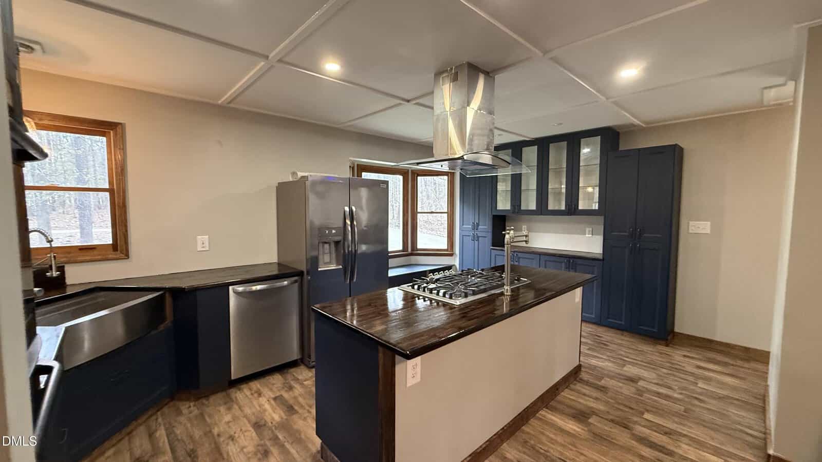 A modern kitchen with wood floors, stainless steel appliances, dark blue lower cabinets, glass-front upper cabinets, a farmhouse sink, center island with a cooktop and hood, and large windows letting in natural light.