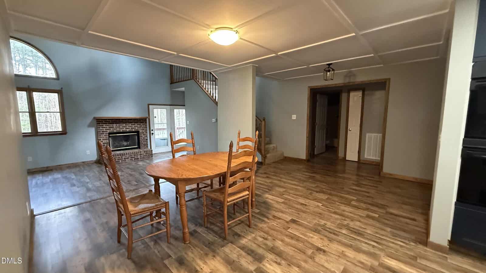 A dining area with a wooden table and four chairs, adjacent to a living room with a brick fireplace, wood floors, gray walls, and a view of stairway and open doorways in the background.