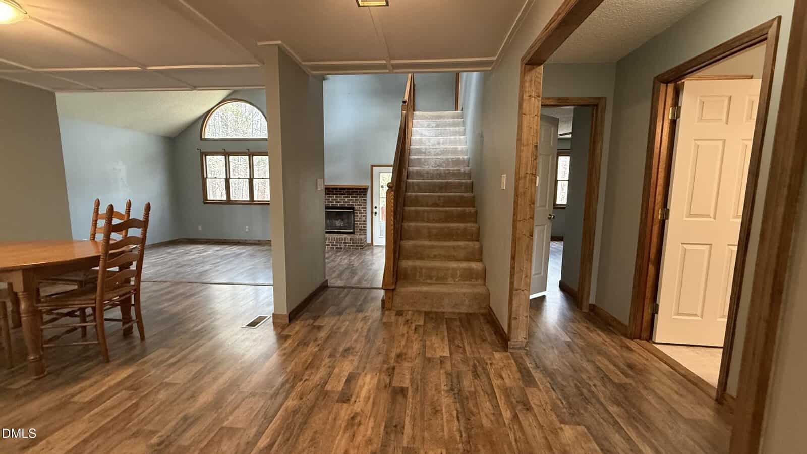 A view of a home's interior featuring a carpeted staircase, a dining area with a wooden table and chairs, wood-look flooring, and multiple doorways, including one leading to a room with a fireplace and large windows.