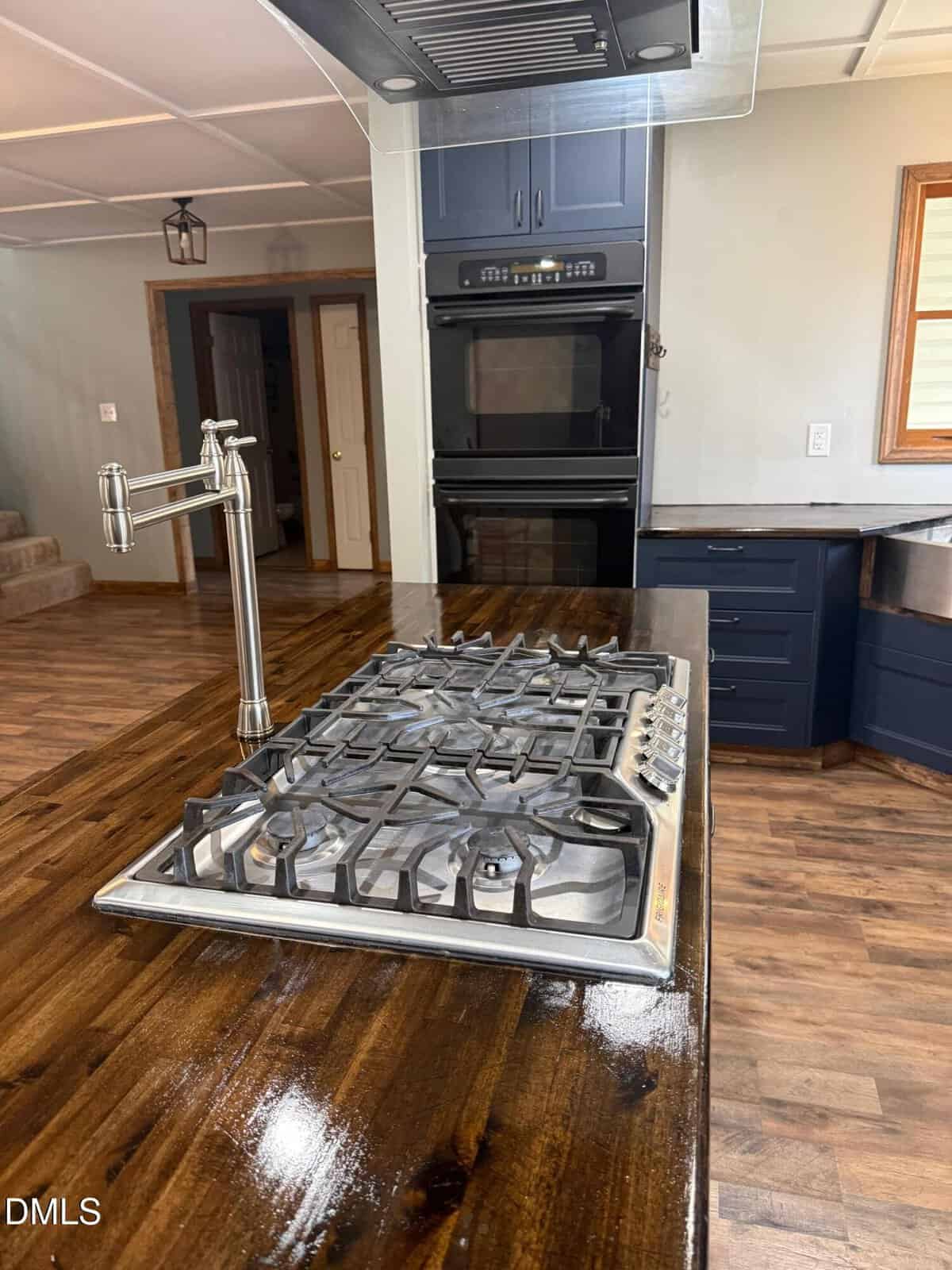 A modern kitchen with a gas cooktop on a wooden island, a pot filler faucet, dark cabinets, double wall ovens, and brown wood floors.