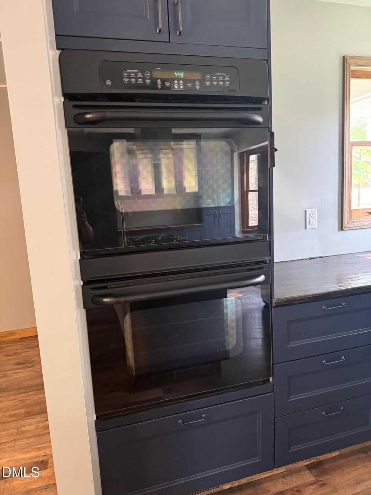 A modern black double wall oven is built into dark cabinetry beside a kitchen counter. Sunlight streams through a window, and the oven’s digital display is lit.