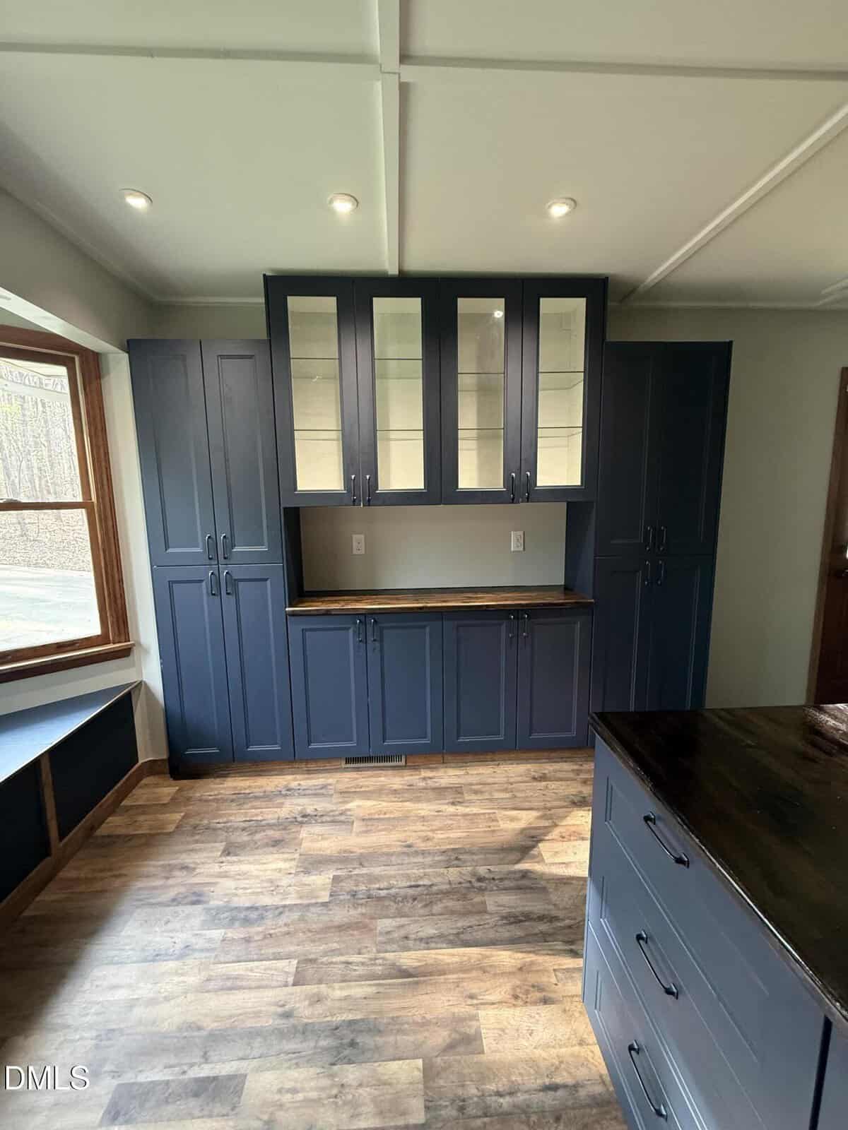 A kitchen with navy blue cabinets, glass-paneled upper doors, wood countertops, recessed lighting, and wood flooring. A window seat is on the left, and an island with dark top is in the foreground.